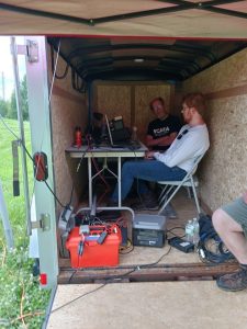 Digital station trailer with two operators, a table with a laptop and transceiver, and various cables running into portable battery packs on the floor