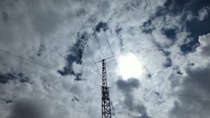 Beam antenna atop an aluminum tower with guy wires visible and the sun and broken clouds above