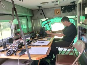 Inside the communications truck. Various equipment is scattered on the fold-down table while an operator adjusts the transceiver